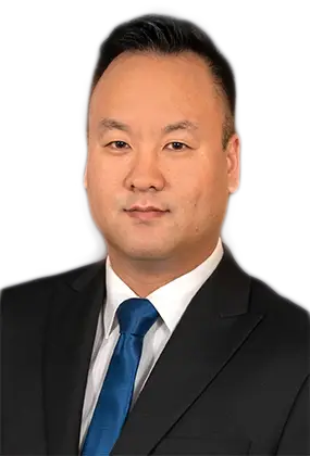 A photograph of Daniel Kim, AS3 Office Manager, is shown on a dark-grey background. Daniel is an Asian male wearing a black suit coat, white dress shirt, and a navy blue tie. He has short black hair and a slightly darker complexion, and is smiling | AS3 Med Spa