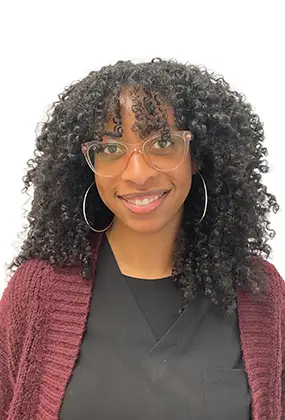 Naia Taylor-Mitchell, front desk receptionist at AS3 Medical Spa, smiling while wearing glasses and black medical scrubs.